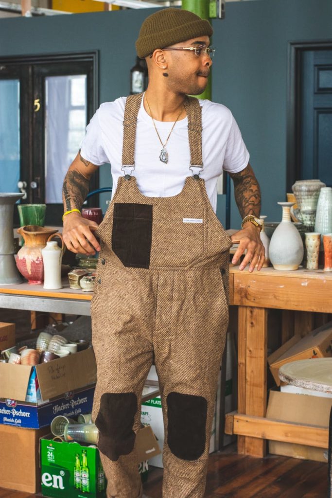 a man wearing dungarees standing in front of a table