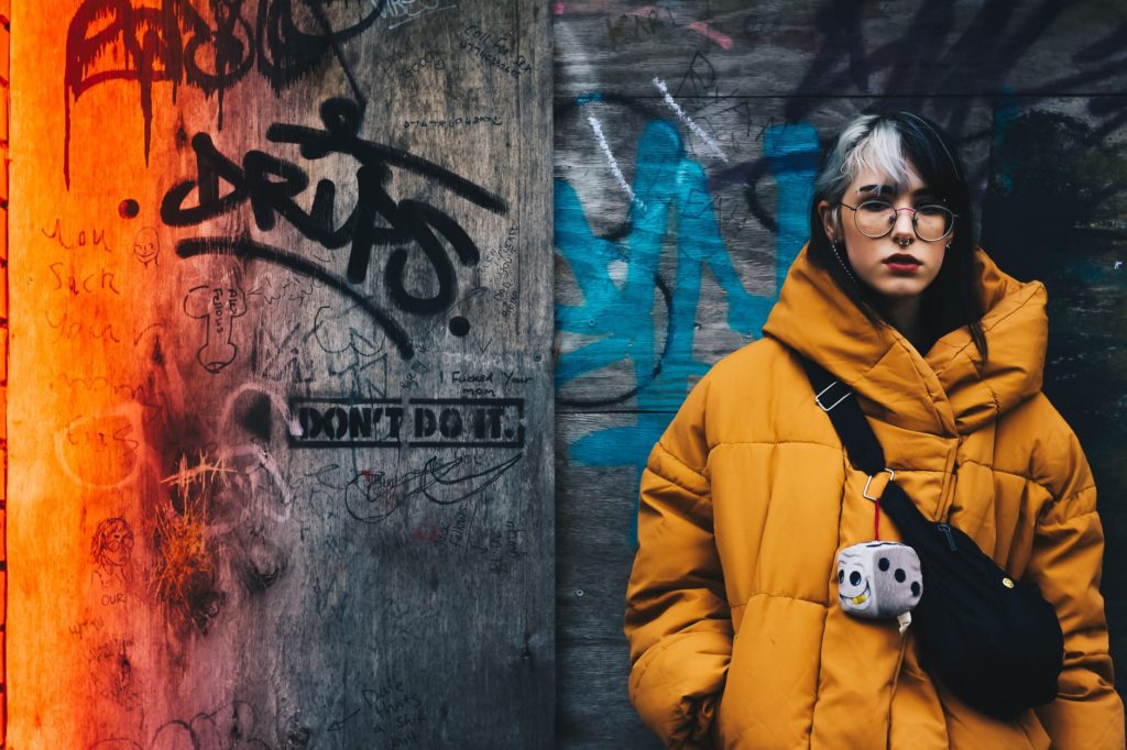 a woman in a mustard quilted jacket with a black crossbody bag and standing in front of a wall with graffiti
