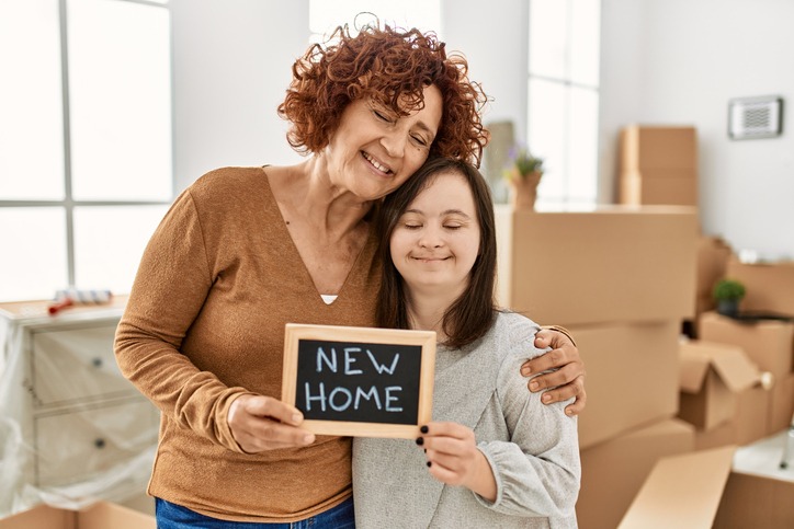 Mature mother and down syndrome daughter moving to a new home, standing by cardboard boxes holding blackboard with new home phrase