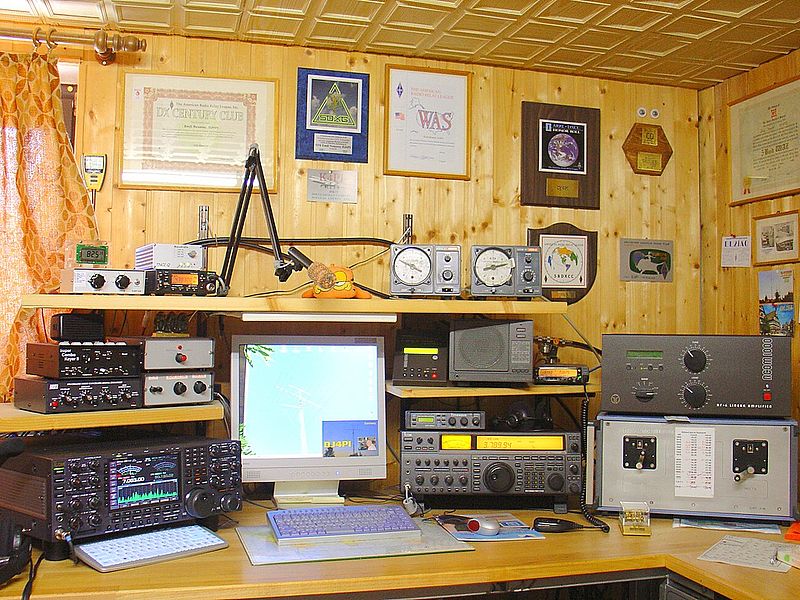 An illustration of an amateur radio station with four transceivers, amplifiers, and a computer for digital modes and logging. Examples of numerous amateur radio prizes, certificates, and QSL cards from international amateur stations are displayed on the wall.