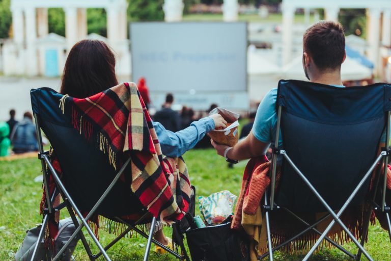 Couple sitting in camp-chairs in city park looking movie outdoors at open air cinema