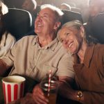 Senior couple, laugh and happy at cinema for movie in crowd, bonding and date with comedy show. Elderly people, smile and excited with fast food, popcorn and watch comic film with love at theater